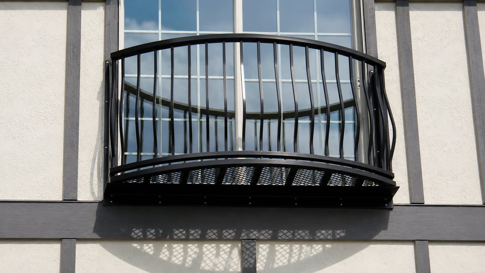 Black aluminum bowed basket-picket balcony with a woven metal floor, mounted beneath an arched window